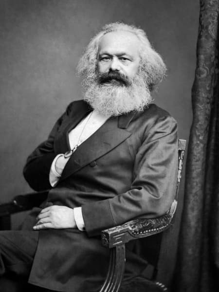 Karl Marx, a bearded man with fuzzy white hair, gazes at the camera with his right hand placed inside his suit jacket.