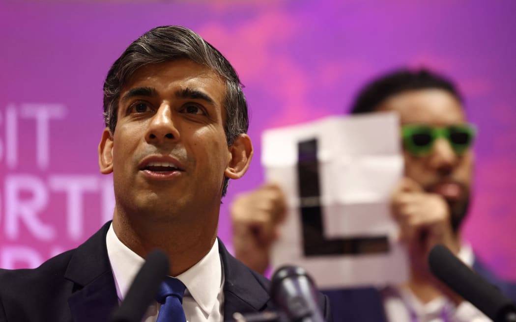 Independent candidate Niko Omilana holds an "L" behind Britain's Prime Minister and Conservative Party leader Rishi Sunak as he delivers a speech after retaining his seat as MP for Richmond and Northallerton in Northallerton, north of England, early on July 5, 2024.