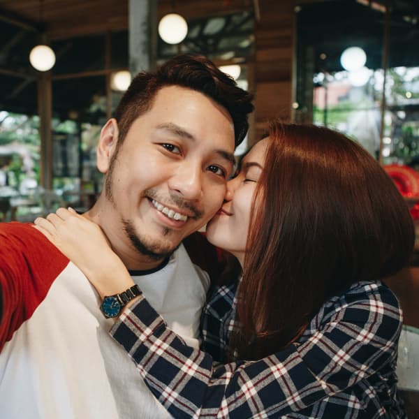 A man smiles for the camera while a woman nuzzles his face.