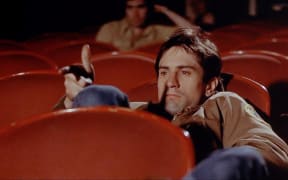 Movie still from Martin Scorsese's film Taxi Driver showing Robert De Niro as Travis Bickle slouching in a cinema with his fingers pointing like a gun barrel.