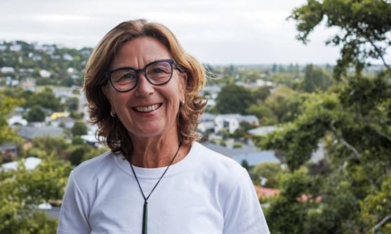 Rebecca Macfie, a middle-aged woman wearing a white top and black glasses, smiles at the camera. She stands outside in front of trees.