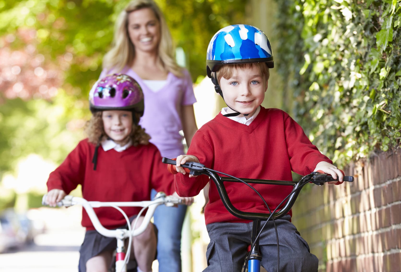 Two children cycle to school with their mother in the background.