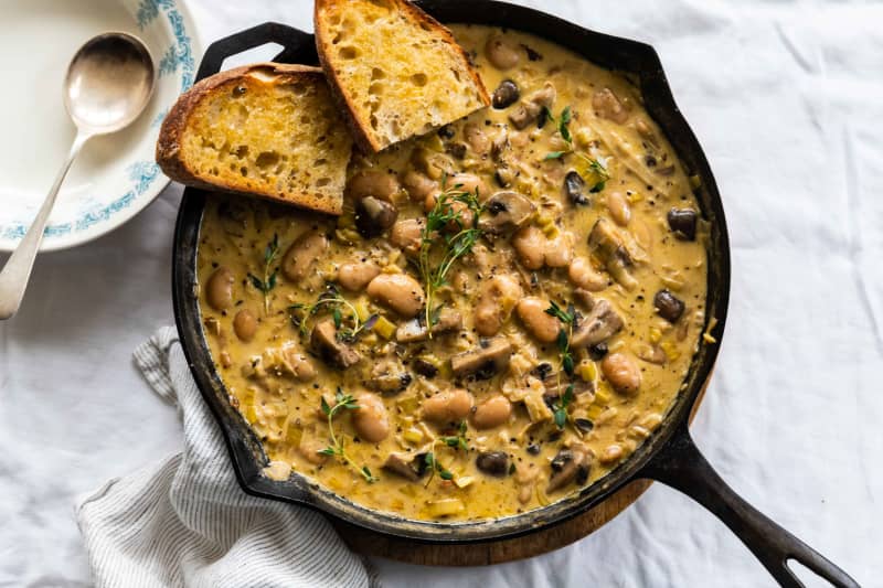A round cast iron frying pan containing a creamy mixture of leeks, mushrooms and butter beans, garnished with two pieces of sliced baguette and sprigs of thyme.