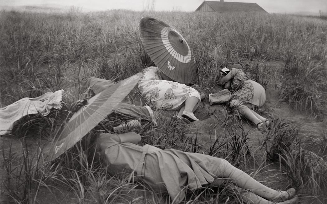 Three women lie on sand dunes, holding parasols over their heads. The photo is old, in black and white.