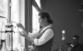 Kate stands in a dimly lit studio by a table of distilling equipment, holding a small glass vial to the light. David sits at a table in the background.