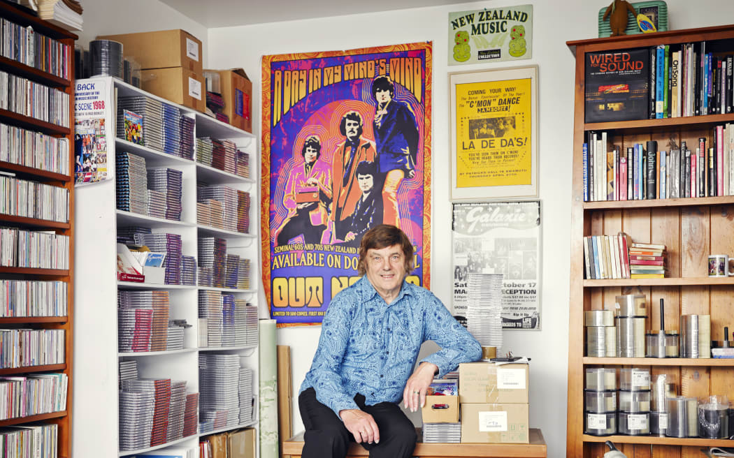 Grant smiles at the camera. He is sitting at a small desk covered in boxes of cassette tapes. The room he is in is covered in colourful music posters and shelves of CDs.