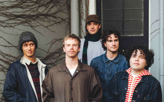 The band is made up of five men. They are stood in front of the door to a house an looknig at the camera.