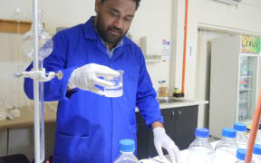 A man in a blue lab coat with white gloves holds up a beaker containing a transparent colourless liquid. He is standing in a lab with glassware and bottles on the lab bench in front of him.
