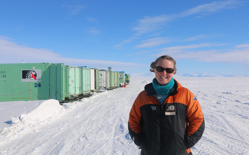 A woman wearing sunglasses, a blue scarf and a black-and-orange warm jacket stands on a flat expanse of white ice beneath a blue sky. On the left, a row of mint-green containers, the closest of which has the Antarctica New Zealand black-and-white fern/penguin logo on it.