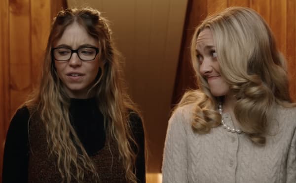 Two young women with long hair stand side by side in front of a wooden wall.