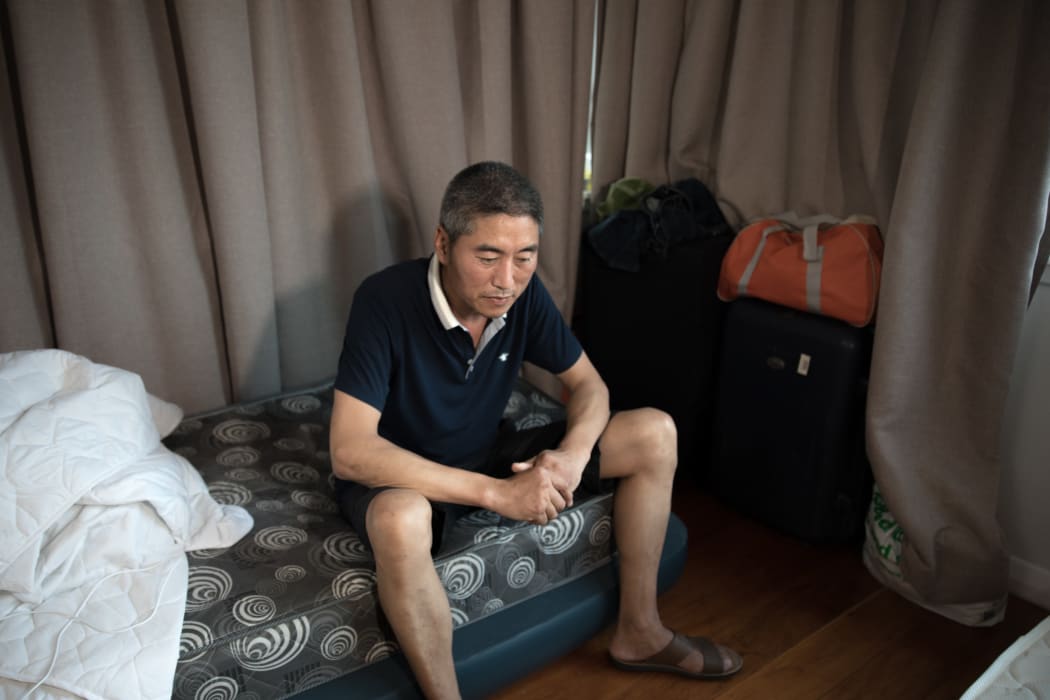 Gao Litong sits on his bed which is a mattress on the floor of a living room which he shares with another Chinese worker who came to New Zealand to work.