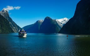 A tourist sightseeing ferry boat travels down the calm, deep blue waters of Milford Sound in the remote Fiordland region in the south west of the South Island of New Zealand