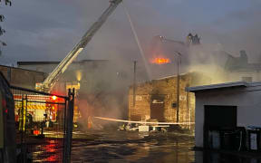 Flames could be seen over the top of a building in Onehunga Mall Road, as firefighters work from ladders, on Sunday night, 25 January, 2026.