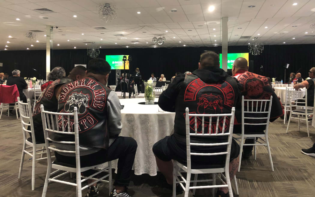 Patched Mongrel Mob members sitting around a table in a conference venue, listening to someone talking on a stage. Their backs are to the camera.
