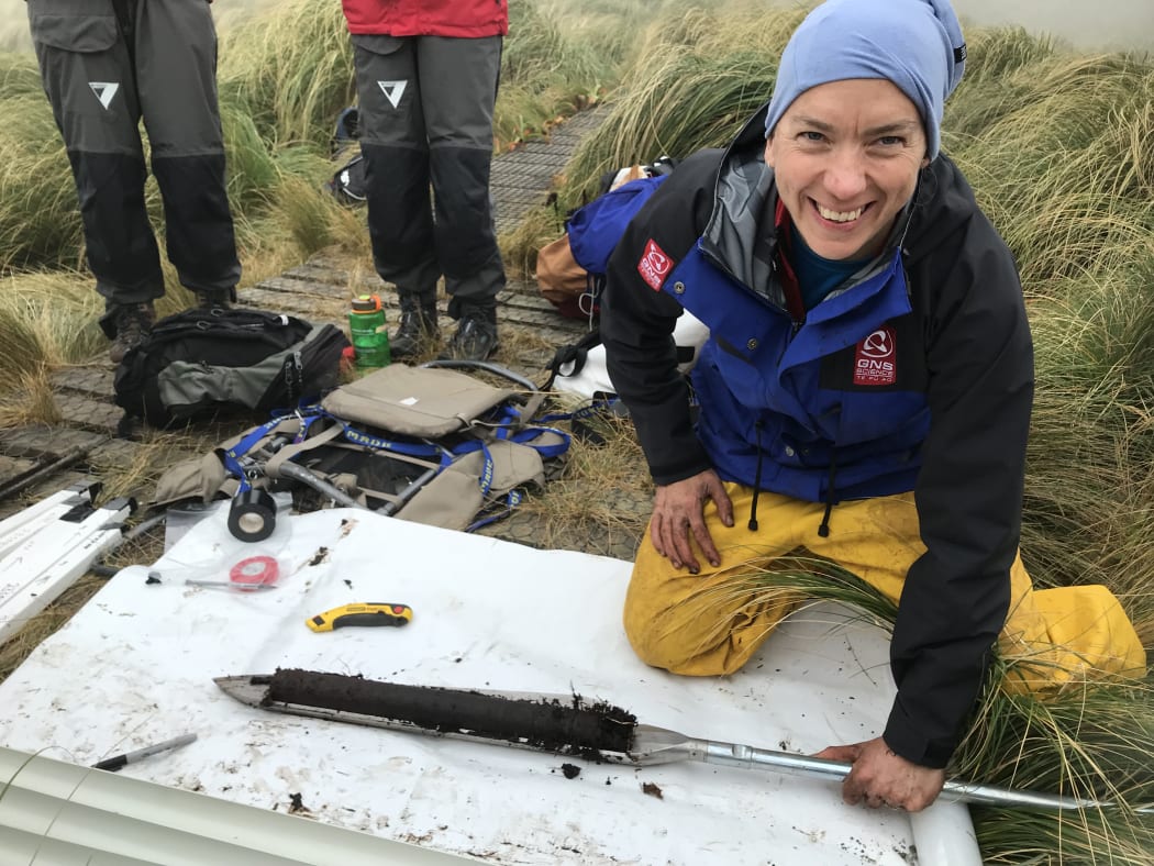 Greer, wearing a GNS Science blue and black jacket, with yellow waterproof pants and a blue bandana on her head is smiling as she holds a corer with a half cylinder core of peat on a white tarp.