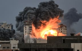 People standing on a rooftop (left), watch as a ball of fire and smoke rises above a building in Gaza City on 7 October, 2023 during an Israeli air strike.
