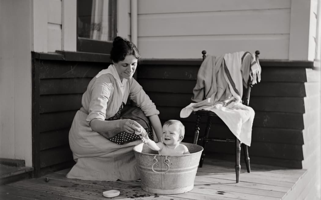 Maud bathes her baby Nancy in a metal tub on the deck of the house. They both smile.