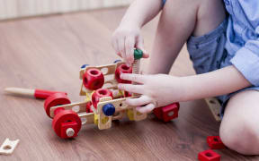the little boy twists the bolt into a wooden machine  on floor in room.