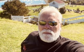 Phil Paikea wears sunglasses and stands in front of a house, a lake, mountains and a blue sky