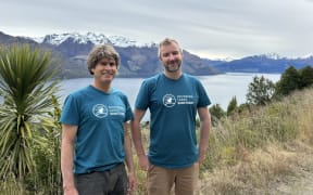 Two men wearing blue t-shirts with 'Southern Lakes Sanctuary' written on them stand next to a spiky tree in front of a lake and snow-capped mountains.