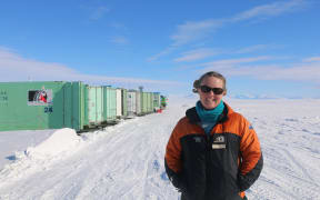 A woman wearing sunglasses, a blue scarf and a black-and-orange warm jacket stands on a flat expanse of white ice beneath a blue sky. On the left, a row of mint-green containers, the closest of which has the Antarctica New Zealand black-and-white fern/penguin logo on it.