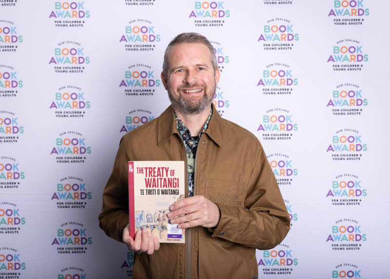 Author Ross Calman, wearing a brown zip up jacket, holds his book, The Treaty of Waitangi.