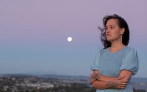 Māori architect Jade Kake wears a blue dress and stands in front of a city scape at sunset with her arms crossed in front of her. The moon can be seen in the background.