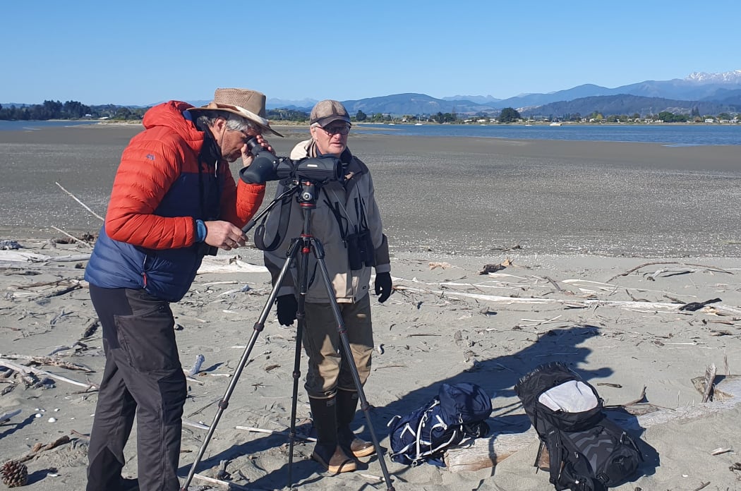 Two men in jackets and hats stand on the sandspit, with driftwood scattered around. One is looking through a large binoculars.