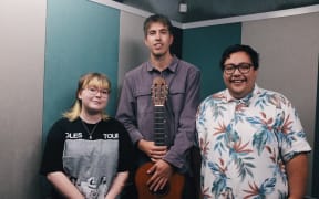 Three people standing in a line together, one person in the middle holding a guitar. They're in a studio.