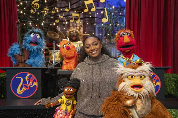 A smiling woman in a grey sweater poses with a group of puppets.