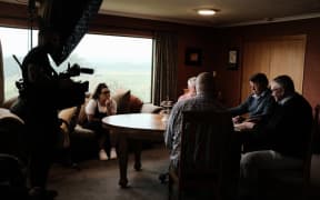 A group of people sat in a dimly-lit living room. A cameraman is operating a camera and microphone. Jenny sits with her chin resting on her fist, listening while Max, Robert, Bill and Stewart sit and talk around a circular wooden table.