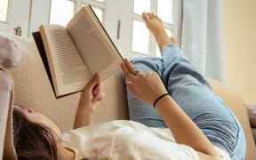 A dark-hared woman lies on the couch reading with her bare feet up.