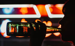 A person in silhouette drinking a bottle of alcohol with bright lights behind them.