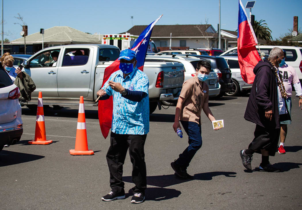 A man waving a Samoan flag and blue face covering dances at the Browns Road netball courts in Manurewa.