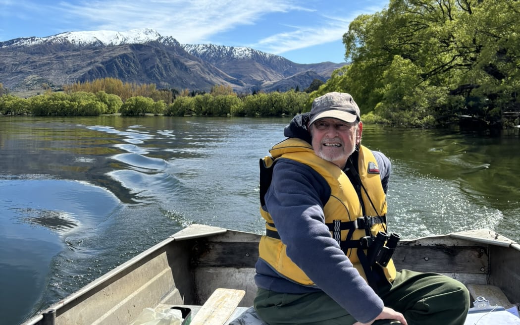 Richard Bowman sits in a metal dinghy (a tinny) on Lake Hayes, wearing a yellow lifejacket and a pair of binoculars around his neck. It's a sunny day and there are snow-dusted mountains in the background.
