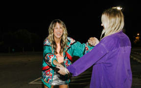 Two blonde women in brightly coloured clothes smile while holding hands