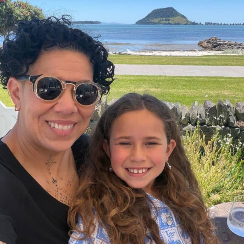A woman with short hair and a child with long hair smile and sit close together with a coastal view behind them.