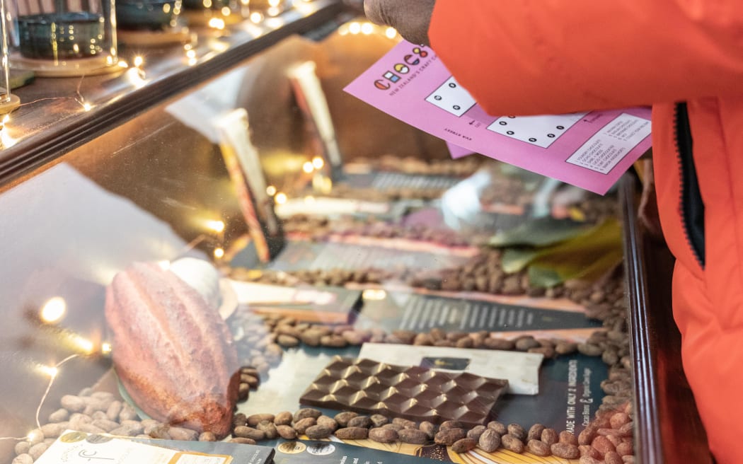 A person leans over a counter full of craft chocolate. The cabinet is lit with golden light, and the displays are surrounded by cocoa beans.