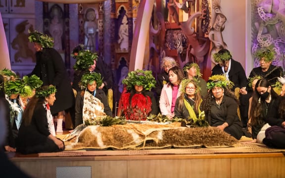 A group of people solemnly gather around kōimi t'chakat, the skeletal remains of Moriori ancestors, which are covered with ceremonial cloaks.