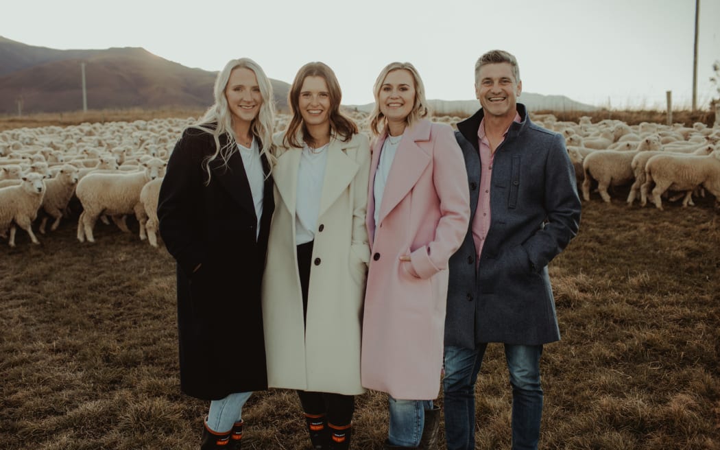 Three women and a man standing in a paddock modelling coats with a flock of sheep in the background