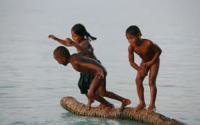 Children in Kiribati play on a partially submerged coconut trunk.
