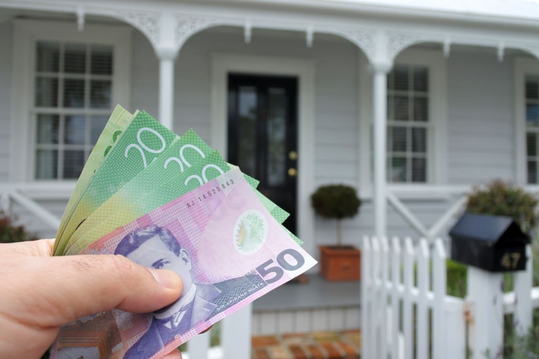 A mans hand holds NZ dollar bills against a front of a traditional villa house in Auckland, New Zealand. Buy, sale, real estate, insurance, mortgage, bank loans and housing market concept.