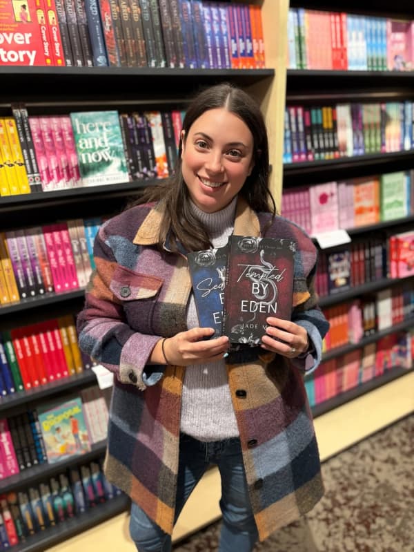 Jade May has long brown hair and stands in front of a bookshelf with a book.