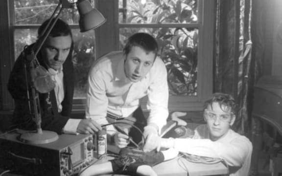 Bressa Creeting Cake are three young men huddled around a desk on which sits a strange-looking machine.