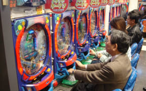 A gentleman, in a Pachinko Parlour, sat at a brightly-coloured Pachinko machine on a blue seat. Others are sat in a line next to him, and are doing the same.
