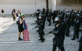 A protester stands holding a U.S. flag while facing a line of law enforcement officers during demonstrations in Los Angeles, California, on June 8, 2025. Following a series of aggressive federal immigration operations in Los Angeles, tensions escalated when Immigration and Customs Enforcement (ICE) agents conducted a high-profile raid on a Home Depot location. The raid sparked widespread protests across the city, where demonstrators decried the targeting of immigrant communities and the separation of families. Clashes soon broke out between protesters and federal agents, prompting U.S. President Donald Trump to deploy 2,000 National Guard troops to the city, a move harshly criticized by California officials, including the governor, as "purposefully inflammatory." (Photo by David Pashaee / Middle East Images via AFP)