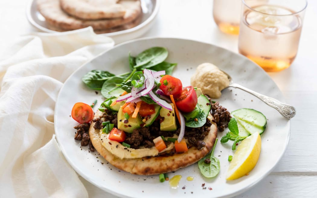Spiced lamb mince with hummus and flatbreads on a round white plate garnished with herbs and a lemon wedge.