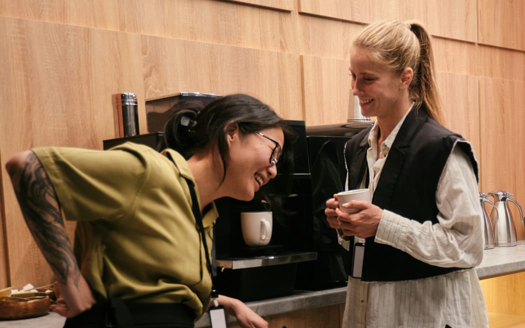 Two women smile next to a coffee machine.