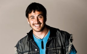 Tammy Davis is a young smiling man wearing a denim vest, blue t-shirt and pounamu pendsnt.