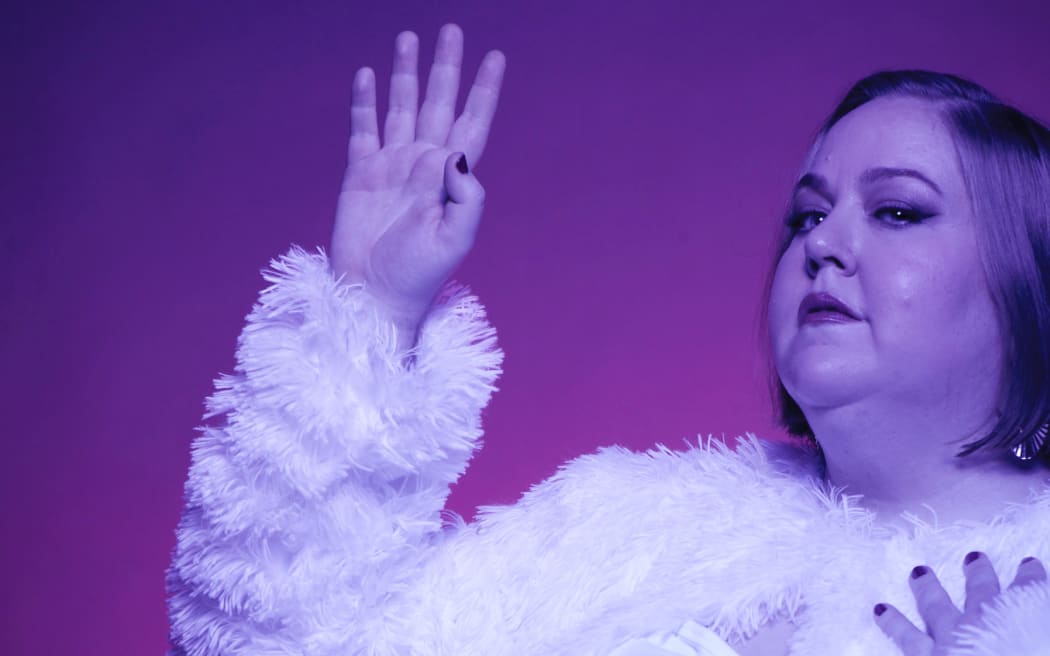 Aubrey Gordon - A fat woman with blonde hair stands unsmiling holding her right hand aloft. She is wearing a striped dress and a fluffy jacket whilst bathed in purple light and set against a pink and purple background.